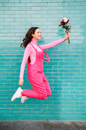 Creative Highlights Symbolbilder Happy woman wearing pink bib overalls jumping with roses in front of blue brick wall model released, Symbolfoto, AMWF02289