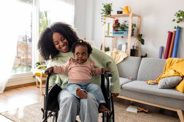 IMAGO Creative, Diversity, Mütter Cute baby girl sitting on young mother with disability in wheelchair at home model released, Symbolfoto property released, JCCMF11739