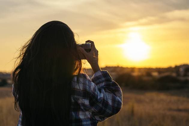 Young woman in checkered shirt taking photos with an analog camera in the field at sunset. The sky is orange and the colors in the image convey tranquility. Model Released PedroMerino_ID19010_565611_025 Copyright: xx