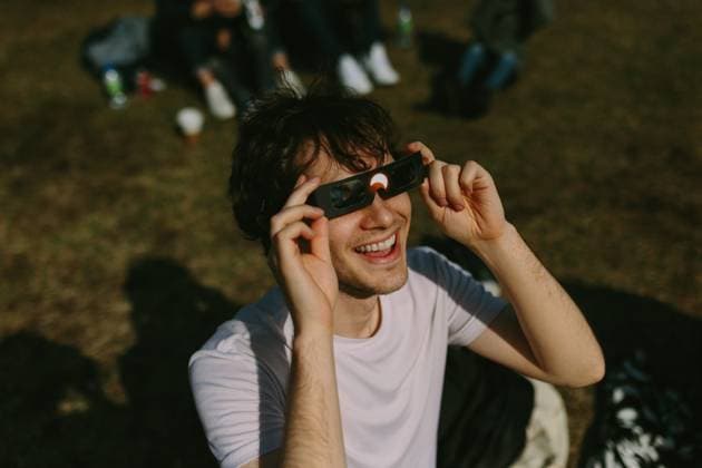IMAGO Creative: Sommer, Sommertage im Stadtpark A cheerful young man enjoys using solar eclipse glasses in a sunlit park, surrounded by a relaxed group of friends casually gathered in the background Model Released CamilleGirard_ID21463_631383_010 Copyright: xCamillexGirardx