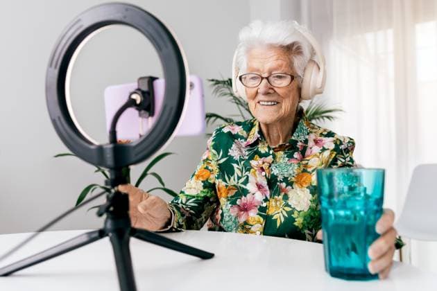 Creative Highlights Symbolbilder An elderly woman in a floral shirt smiles engagingly while recording a podcast She wears headphones and speaks into a microphone, illuminated by a ring light, showcasing modern tech engagement Model Released SergioVictorVega_ID21565_633363_009 Copyright: xSergioxVictorxVegax