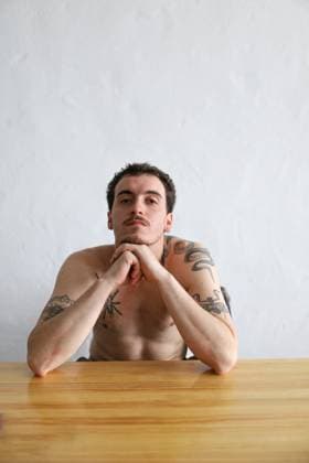 Creative Highlights Symbolbilder A young adult male with intricate tattoos on his arms and torso poses shirtless, sitting at a wooden table, gazing thoughtfully at the camera against a plain white background Model Released Gaorstudio_ID22007_644354_004 Copyright: xGaorstudiox