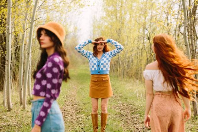 Three stylish women walk through a sunlit forest, surrounded by budding trees. The central figure, wearing a blue and white checkered sweater, stands out as she scouts the path ahead Model Released CamilleGirard_ID21737_639678_028 Copyright: xCamillexGirardx