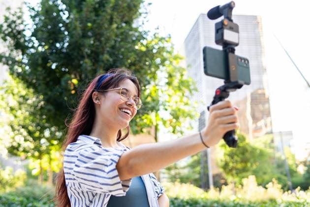 IMAGO Creative, Lifestyle, Frühling in der Stadt A cheerful young woman records herself using a smartphone attached to a gimbal, capturing playful moments while in a sunny urban park Model Released DavidMunoz_ID22797_663732_022 Copyright: xDavidxMunozx