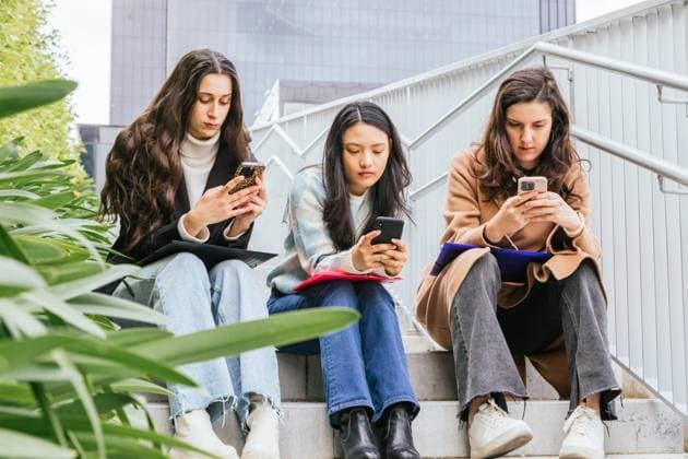 Three young female friends sit closely on outdoor stairs, each deeply engaged in her smartphone, surrounded by city buildings Model Released IgnacioFerrandiz_ID22609_659648_010 Copyright: xIgnacioxFerrandizx RECORD DATE NOT STATED