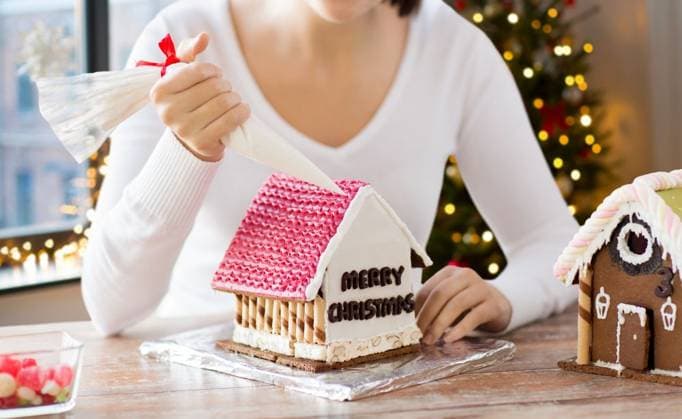 cooking, holidays and people concept - woman with pastry bag making gingerbread house at home over christmas tree lights background. woman making gingerbread house on christmas ,model released, Symbolfoto