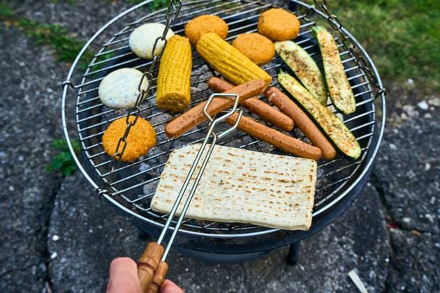 Bavaria, Germany - 26 July 2024: A colorful mix of vegetarian delicacies such as corn on the cob, vegan sausages, zucchini and grilled cheese sizzle on a charcoal grill. A man turns a meatless sausage with a pair of tongs *** Ein bunter Mix aus vegetarischen Köstlichkeiten wie Maiskolben, vegane Würstchen, Zucchini und Grillkäse brutzeln auf einem Kohlegrill. Ein Mann wendet mit einer Zange eine fleischlose Bratwurst