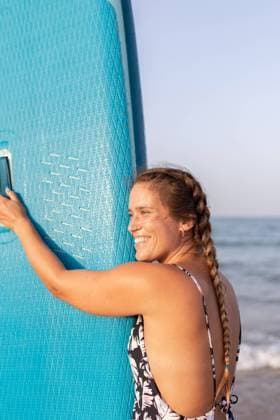 Creative Highlights Symbolbilder MR_GloriaBanuls_PadelSurf.jpg Side view of wet happy female surfer standing with blue SUP board on sandy seashore in summer and looking away Model Released LeoPastor_ID18063_543417_007 Copyright: xLeoxPastorx