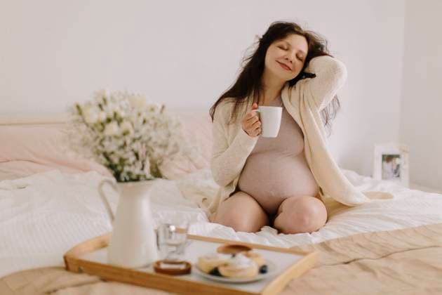 Creative Highlights Symbolbilder Smiling pregnant woman holding coffee mug sitting on bed at home model released, Symbolfoto property released, MCHF00127