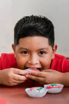 Creative Highlights Symbolfotos A joyful young boy in a vibrant red shirt delightfully eats festive Christmas cupcakes with colorful frosting, capturing the excitement and happiness of holiday treats Model Released PatBueno_ID24493_709899_004 Copyright: xPatxBuenox