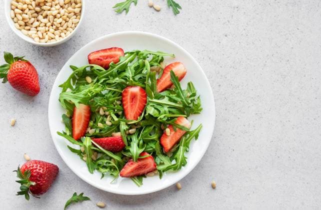 A vibrant plate of strawberry arugula salad, garnished with toasted pine nuts, presented on a white plate, set against a light grey backdrop. The freshness of the greens and the ripeness of the strawberries suggest a healthy, nourishing meal option. Model Released OlenaRudo_ID23469_682014_024 Copyright: xx