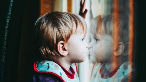 Creative Highlights Symbolfotos A young child with blonde hair looking out a window, reflecting on the glass, wearing a colorful bib. 25765449 ,model released, Symbolfoto RECORD DATE NOT STATED