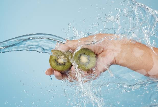 Hands, beauty and kiwi in a studio with splash for skincare, shower and organic treatment against a blue background. Hand, water and fruit with woman wellness model grooming, hygiene and cleaning. model released property released Copyright: xwww.peopleimages.comx