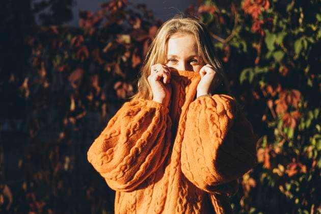 A woman wearing a cozy knitted orange sweater, partially covering her face, stands amidst vibrant autumn foliage Model Released HannaYakimenko_ID25074_726418_025 Copyright: xHannaxYakimenkox