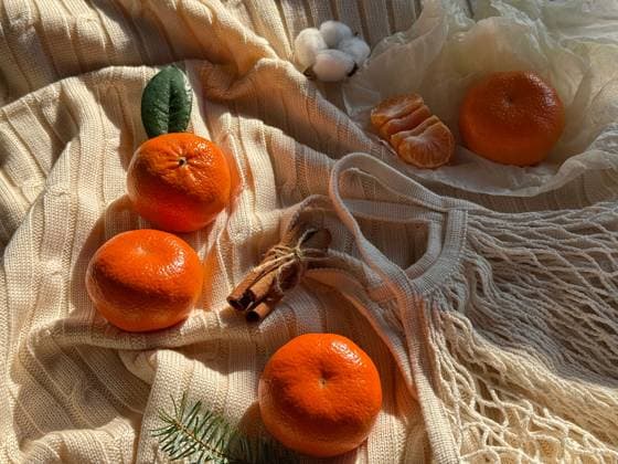 Top view of fresh mandarins on a textured beige fabric with natural sunlight. Includes a cotton puff, peeled segment, mesh produce bag, and cinnamon sticks, conveying a cozy, homely vibe. Model Released IrinaChetverikova_ID24791_718545_012 Copyright: xIrinaxChetverikovax RECORD DATE NOT STATED