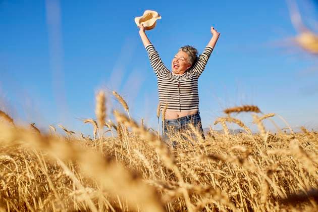 Cheerful mature woman with bucket hat jumping amidst wheat crops in field model released, Symbolfoto property released, ESTF00185