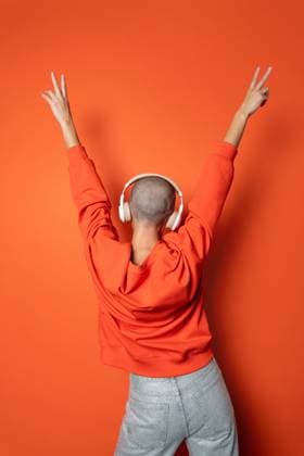 Creative Highlights Symbolbilder 783449156 Bald person in a vibrant orange sweatshirt and white headphones caught in a jubilant pose with arms raised, on a solid orange background, expressing joy and liberation through music. Model Released ManuelRuiz_ID25519_737680_048 Copyright: xManuelxRuizx
