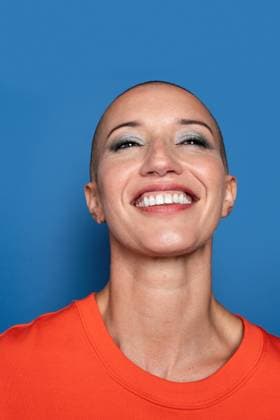Creative Highlights Symbolbilder 783449156 Portrait of a joyful bald Caucasian adult woman wearing an orange t-shirt. She displays a bright, toothy smile against a vibrant blue background, showcasing confidence and happiness. Model Released ManuelRuiz_ID25519_737692_060 Copyright: xManuelxRuizx