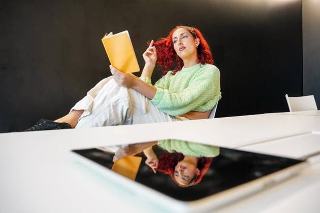 Creative Highlights Symbolbilder 783449156 A trans woman with red hair relaxes in a modern work space, reflecting deeply while reading a book Her thoughtful expression conveys contemplation and pride Model Released SheilaAlonso_ID24681_715021_027 Copyright: xSheilaxAlonsox RECORD DATE NOT STATED