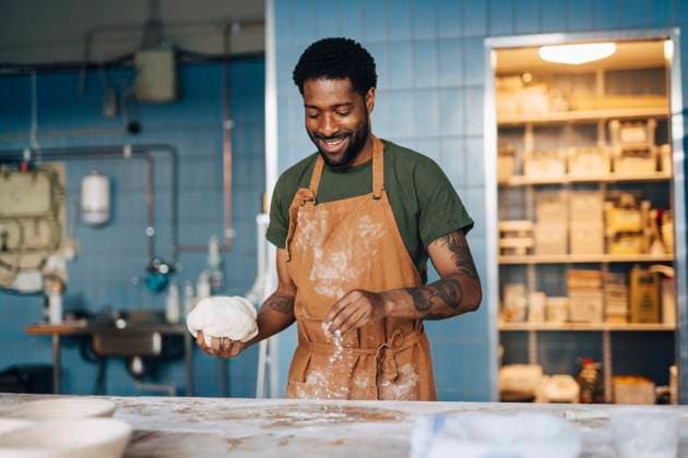 Smiling male baker dusting flour on table and holding dough while working at bakery Sweden, Stockholm, model released, property released Copyright: xMaskotx , MA114843 MA114843