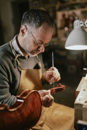 Italienischer Geigenbauer mit Brille und Schürze, der in einer schwach beleuchteten Werkstatt, die von einer fokussierten Lampe beleuchtet wird, akribisch Lack auf eine Geigenschnecke aufträgt Italian violin maker, wearing glasses and an apron, meticulously applying varnish to a violin scroll in a dimly lit workshop illuminated by a focused lamp Copyright: imageBROKER Columbo ibxaug11935958.jpg ,model released, Symbolfoto ,property released