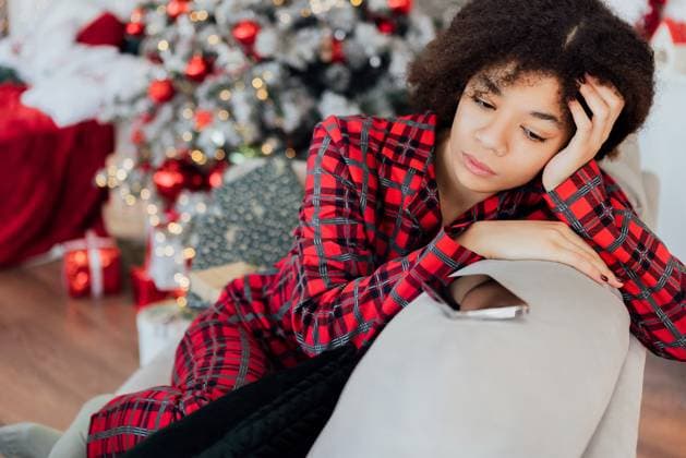 Junge Afrikanerin im Pyjama sitzt auf der Couch und schaut nachdenklich auf ihr Telefon. Das schwarze Mädchen ist traurig und wartet auf einen Anruf. Young African in pajamas is sitting on couch and thoughtfully looking at her phone. Black girl is sad and waiting for call. Sorrowful woman in living room with festive Christmas decorations and gifts ,model released, Symbolfoto Copyright: xZoonar.com DashaxPetrenkox 22526948 ,model released, Symbolfoto ,property released
