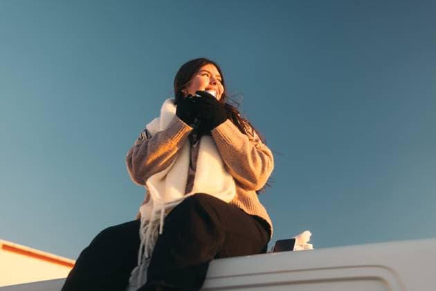 Creative Highlights Symbolbilder Low angle of a cheerful young Latina model from Colombia is captured basking in the golden sunlight of Iceland. Perched atop a vehicle, she gazes skyward with a joyful expression, clad in a cozy sweater and scarf. Model Released Centric_ID25620_739906_016 Copyright: xCentricx RECORD DATE NOT STATED