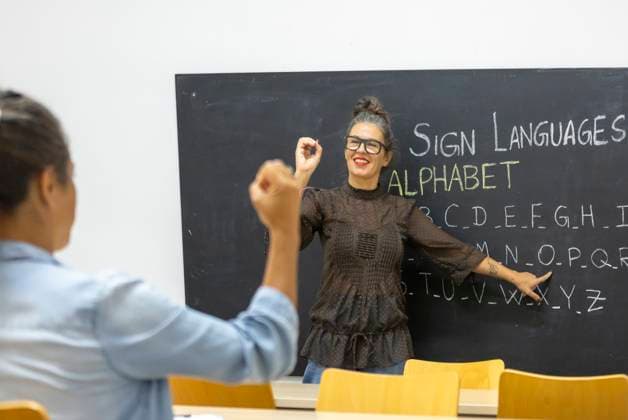 An instructor teaches sign language, pointing to an alphabet chart on a chalkboard A student practices hand signs, fostering communication skills and inclusivity Model Released JuanAizpuru_ID25565_738854_002 Copyright: xJuanxAizpurux