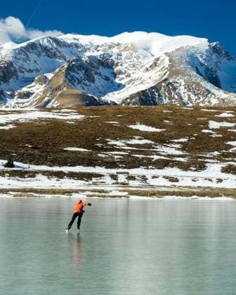 IMAGO Creative, Winter, Auf dem Land A person ice skating on a frozen lake with snow-covered mountains in the background under a clear blue sky. 28391770 RECORD DATE NOT STATED
