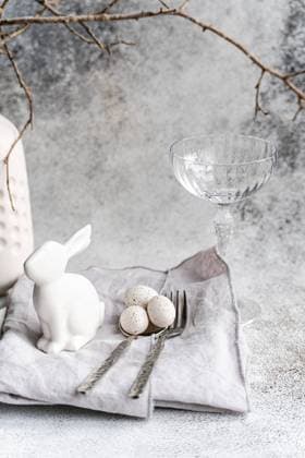 An elegant Easter-themed display featuring a white ceramic rabbit, three speckled eggs, and a clear glass. This setup is complemented by silverware and a textured napkin, presenting a serene holiday mood. Model Released AnnaBogush_ID26004_750181_015 Copyright: xAnnaxBogushx RECORD DATE NOT STATED