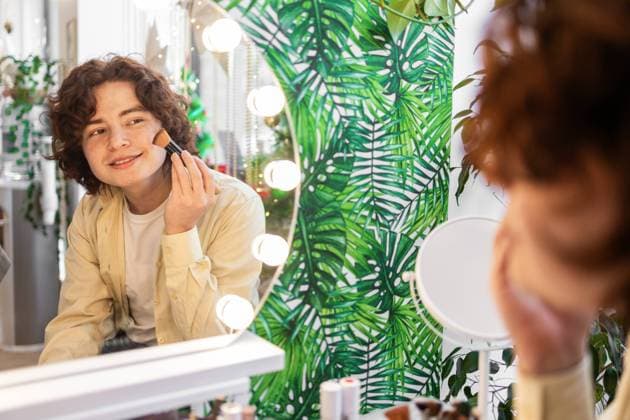 IMAGO Creative, Naturkosmetik A young man with curly hair smiles while applying organic skin care with a brush, standing in front of a mirror surrounded by bright lights and vibrant green leaf decor Model Released DavidMunoz_ID26067_751984_048 Copyright: xDavidxMunozx