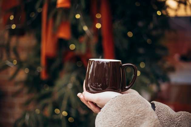 Cropped unrecognizable young woman holding a warm coffee mug in front of a beautifully decorated Christmas tree, capturing the essence of comfort and warmth during the winter season. Model Released HannaYakimenko_ID26106_753104_019 Copyright: xHannaxYakimenkox