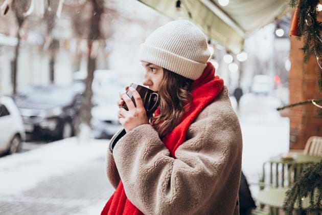 IMAGO Creative, Winter, In der Stadt A young thoughtful woman is pictured standing outdoors on a snowy winter day, sipping from a mug. She is dressed in a warm beige coat, white knit beanie, and a vibrant red scarf, with soft snowflakes drifting around her. Model Released HannaYakimenko_ID26106_753113_025 Copyright: xHannaxYakimenkox