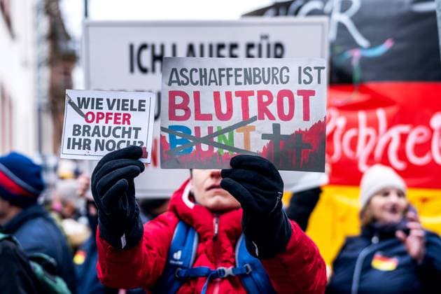 Aschaffenburg, Bavaria, Germany - January 26, 2025: Demonstration and march after knife attack against current government and political actions. A demonstrator holds signs with the inscription: Aschaffenburg is blood red instead of colorful and How many more victims do you need? in the middle of a rally. The message is socio-politically polarized *** Demonstration und Marsch nach Messerattacke gegen aktuelle Regierung und politisches Handeln. Ein Demonstrierender hält Schilder mit der Aufschrift: Aschaffenburg ist blutrot statt bunt und Wie viele Opfer braucht ihr noch? inmitten einer Kundgebung. Die Botschaft ist gesellschaftspolitisch polarisiert