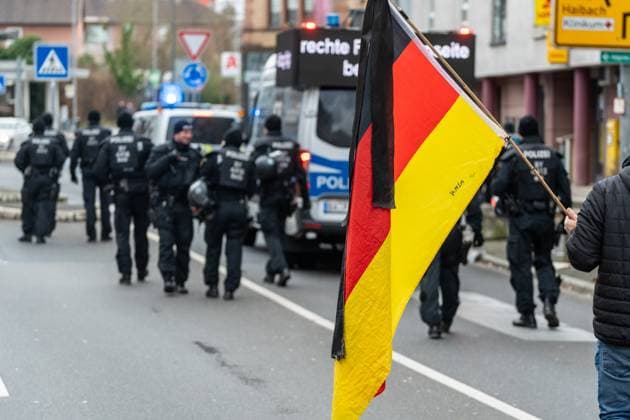 Aschaffenburg, Bavaria, Germany - January 26, 2025: A group of police officers in protective clothing move towards an emergency vehicle. In the foreground is a person with a German flag, which is symbolically carried during the demonstration in Aschaffenburg. *** Eine Gruppe von Polizisten in Schutzkleidung bewegt sich in Richtung eines Einsatzfahrzeugs. Im Vordergrund ist eine Person mit einer Deutschlandfahne zu sehen, die symbolisch während der Demonstration in Aschaffenburg getragen wird.