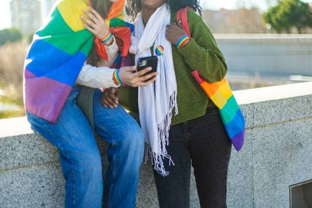 IMAGO Creative, Lifestyle, Frühling in der Stadt Happy lesbian couple using mobile phone and showing rainbow flag Two women smiling and using a smartphone, proudly displaying their support for lgbtq plus rights in a vibrant urban setting Model Released SloomStudio_ID26490_764424_064 Copyright: xcristinaderojasfotografiax