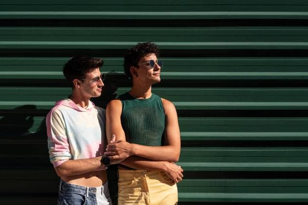 IMAGO Creative, Diversity, Teenager: LGBTQIA+ A joyful gay couple stands together, smiling in the sunlight against a green wall. They exude a sense of happiness and love, enjoying their moment in a casual setting. Model Released ManuelRuiz_ID26527_766006_084 Copyright: xManuelxRuizx