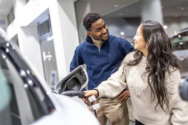 Lächelndes Paar, das die Ladestation eines Elektrofahrzeugs untersucht, während es sich in einem Autohaus über die Möglichkeiten eines nachhaltigen Lebens informiert Smiling couple examining the charging socket of an electric vehicle while exploring options at a car dealership, embracing sustainable living Copyright: imageBROKER UnaixHuizi ibxuhu12460369.jpg ,model released, Symbolfoto ,property released