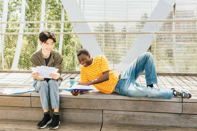 Two friends study outdoors on a sunny day, sitting and laying on a wooden steps surrounded by greenery, engaged in discussion. One holds documents, the other a folder. Model Released IgnacioFerrandiz_ID26810_773769_008 Copyright: xIgnacioxFerrandizx RECORD DATE NOT STATED