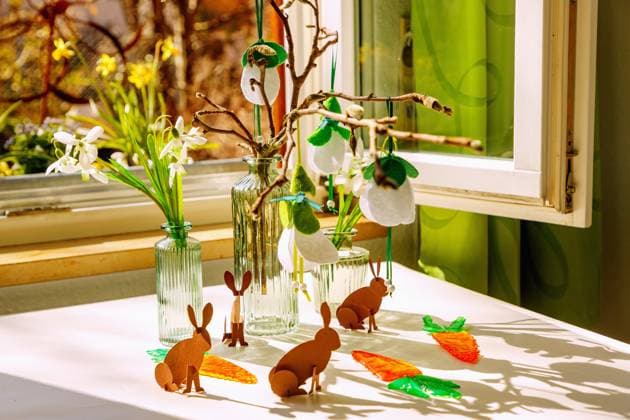 Osterdeko mit Papier-Osterhasen und Karotten aus Schmelzgranulat, Glasvasen mit Schneeglöckchen aus Plastik und Zweigen mit Schneeglöckchen aus Filz auf einem weißen Tisch mit Blick durch geöffnetes Fenster auf Garten und blühende Narzissen *** Easter decoration with Paper Easter bunnies and Carrots from Melting granulate, Glass vases with Snowdrops from Plastic and Branches with Snowdrops from Felt at a white Table with View through Geöffnetes Window at Garden and Flowering Daffodils