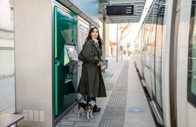Creative Highlights Symbolfotos A woman with a small chihuahua waits near a tram station, standing next to a ticket machine. The urban setting captures modern city life and public transport ambiance. Model Released DavidMunoz_ID27485_789475_004 Copyright: xDavidxMunozx