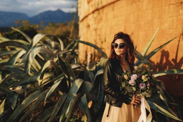 Die Braut in einem schönen pastellfarbenen Kleid und mit Sonnenbrille hält einen Blumenstrauß in den Händen vor dem Hintergrund von Agavensträuchern und der Burgmauer Bride in a beautiful pastel dress and sunglasses holds a bouquet of flowers in her hands against the background of agave bushes and the castle wall. Copyright: imageBROKER NadtochiyxVladimir ibxiqx13172695.jpg ,model released, Symbolfoto ,property released