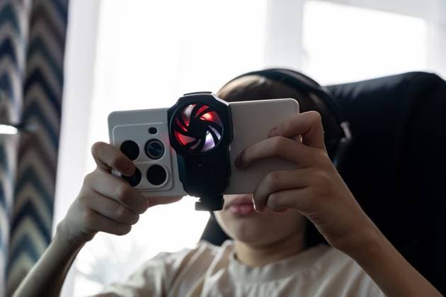 IMAGO Creative, Lifestyle, Gaming A teen boy plays on a smartphone equipped with an external cooling fan attachment while wearing headphones. The setup reflects immersive mobile gaming in a tech-savvy, indoor environment Model Released Ikvyatkovskaya_ID28180_830161_028 Copyright: xIkvyatkovskayax RECORD DATE NOT STATED
