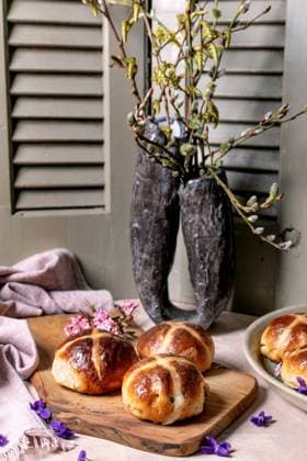 IMAGO Creative, Ostern, Bräuche, Osterbrunch Traditionelle Osterbrötchen im Teig Hot cross buns on wooden cutting board with spring decoration. Easter pastry styled with willow branches in rustic vase, pink blossoms and violet flowers against vintage shutters background. (License=RF) Copyright: xZoonar.com NATASHAxBREENx 23381619