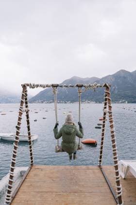 Creative Highlight Symbolbilder Mädchen in Jacke und Hut schaukelt auf einer Schaukel am Pier und blickt auf das Meer und die Berge. Rückansicht Girl in a jacket and hat swings on a swing on the pier and looks at the sea and mountains. Back view. Copyright: imageBROKER NadtochiyxVladimir ibxiqx13181466.jpg ,model released, Symbolfoto ,property released