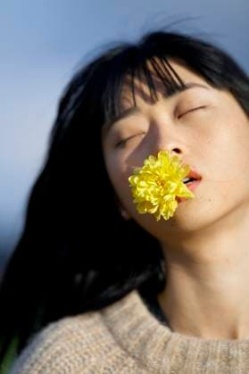 Creative Highlights Symbolbilder Woman with closed eyes holds a yellow flower in her mouth against a blurred blue background. Brandenburg, Germany Berlin Brandenburg Germany bola08130 Copyright: xConnectxImagesx ,model released