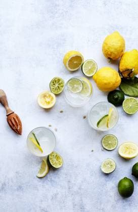 Top view of refreshing summer drinks with slices of lemon and lime on a textured white background. Wood citrus squeezer and scattered seeds add rustic charm. Model Released GeorgiaGold_ID28580_843685_002 Copyright: xGeorgiaxGoldx