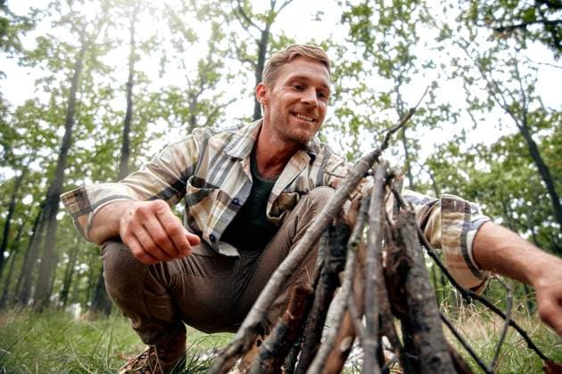 Joyful Outdoorsman Getting Ready to Prepare Campfire in a Beautiful Lush Forest Setting A cheerful man with a warm smile crouches by a flickering campfire, enjoying its warmth and the beauty of the forest, embracing outdoor adventures and reflecting on lifes simple pleasures Model Released OrionProduction_ID27801_798220_052 Copyright: xTonyBeardyx