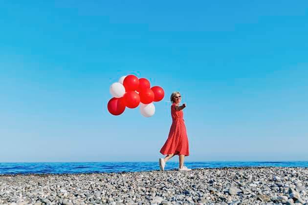 Blond woman in red polka dot dress holding balloons on a pebble beach by the Ionian Sea on a sunny day. model released, Symbolfoto, CATF00697