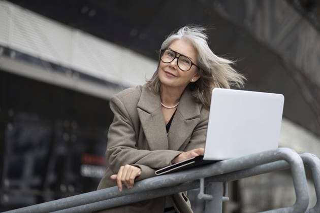 Eine selbstbewusste Geschäftsfrau mittleren Alters mit grauen Haaren und Brille arbeitet mit ihrem Laptop, während sie sich an einem sonnigen Tag an ein Geländer in einer modernen städtischen Umgebung lehnt A confident middle-aged businesswoman with grey hair and glasses engages with her laptop while leaning against a railing in a contemporary urban location on a sunny day Copyright: imageBROKER IvanxTraimak ibxiqx13795691.jpg ,model released, Symbolfoto ,property released